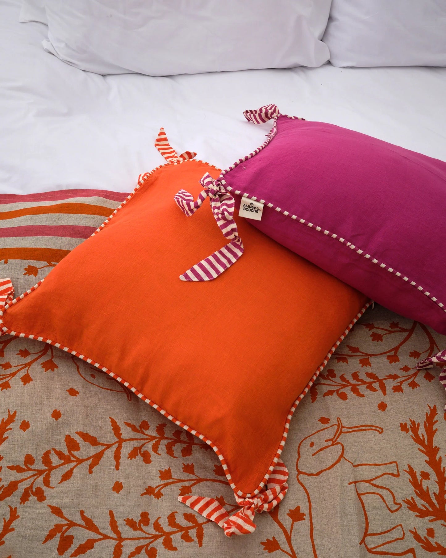 Two decorative Amuse la Bouche cushions one orange and one Fuchsia, both edged with striped piping and bows.
Feather inner included.
