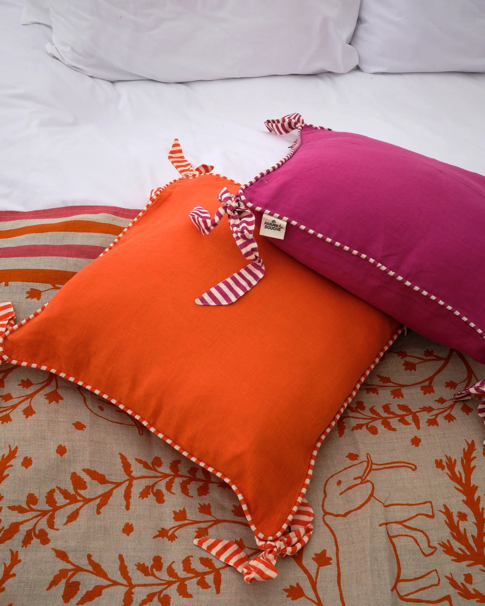 Two decorative Amuse la Bouche cushions one orange and one Fuchsia, both edged with striped piping and bows.
Feather inner included.